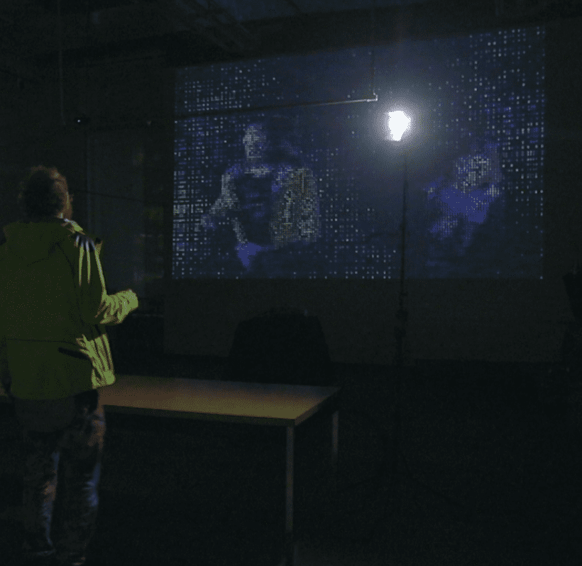 In a dark room, a person stands across from a large video projection. The projected video mirrors the room and the people within it. The images in the video are made up of ascii symbols.