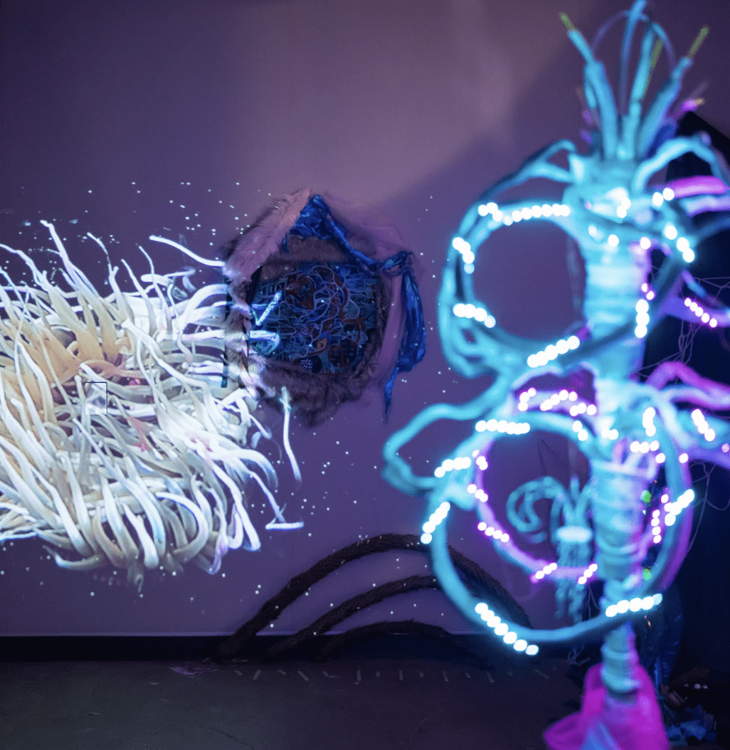 A large, brightly-lit, branching structure stands to the right of the image, in a room filled with purple light. On the wall behind the sculpture is a projection of abstracted anemone. The video overlays an oil painting of twisting shapes. The oil painting is framed with loose, fluffy cloth. Against the wall and on the ground is an arrangement of tiles and monkey tree branches.