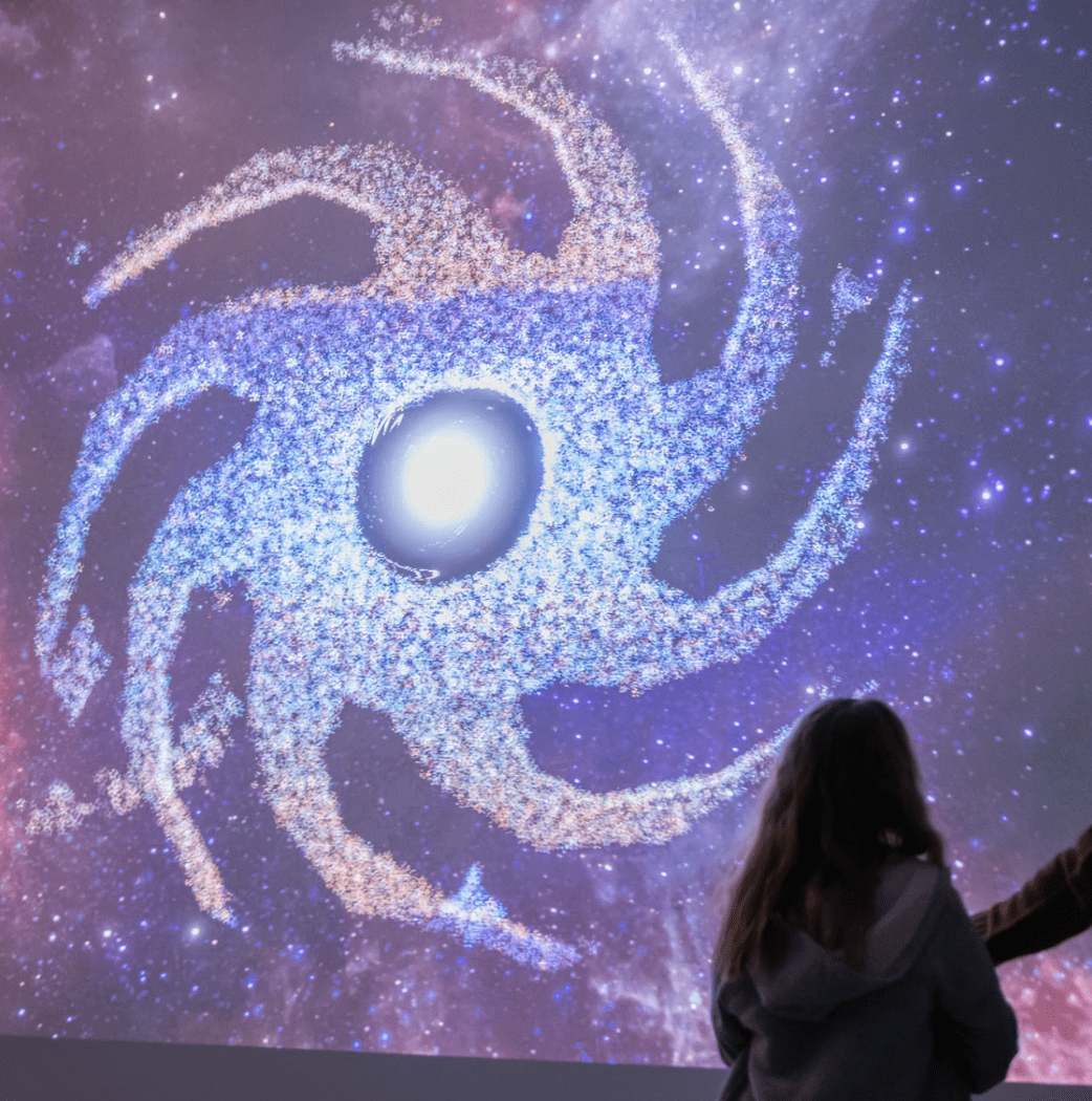 A large swirling shape made of particles is projected onto a wall. A figure stands in front of the projection, to the right of the scene.