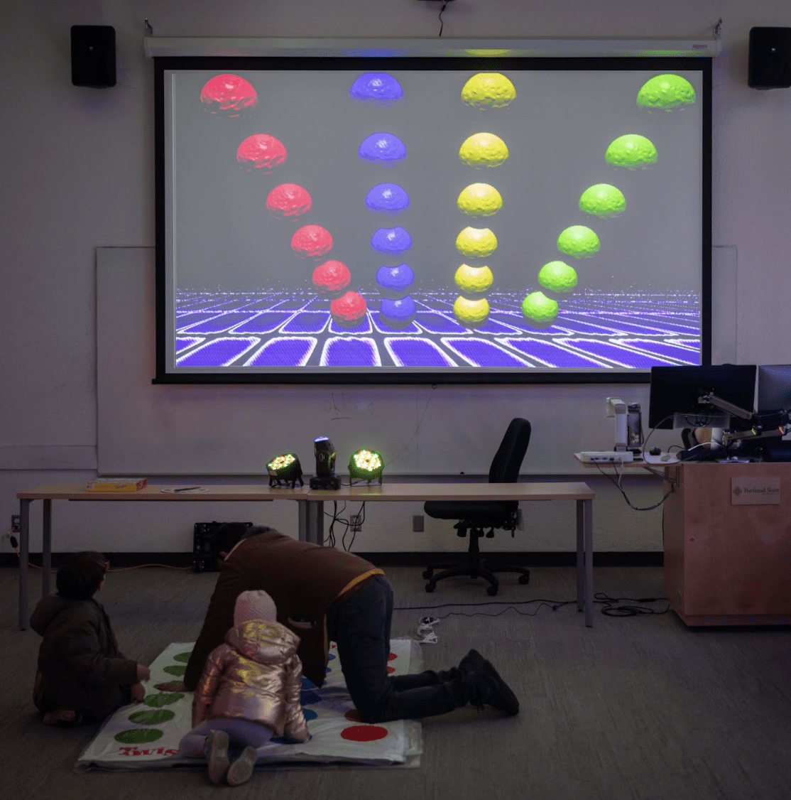 An abstract video featuring rows of circles is displayed onto a classroom projector screen. A twister game is set up on the ground in front of the video. Three people are interacting with the twister game. (Each of the twister circles triggered sounds).