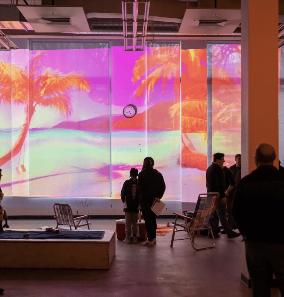 A group of people look up at a large video projection. The video depicts a beach scene, with colors shifted into hues of pink and orange. Beach chairs are set up in front of the projection.