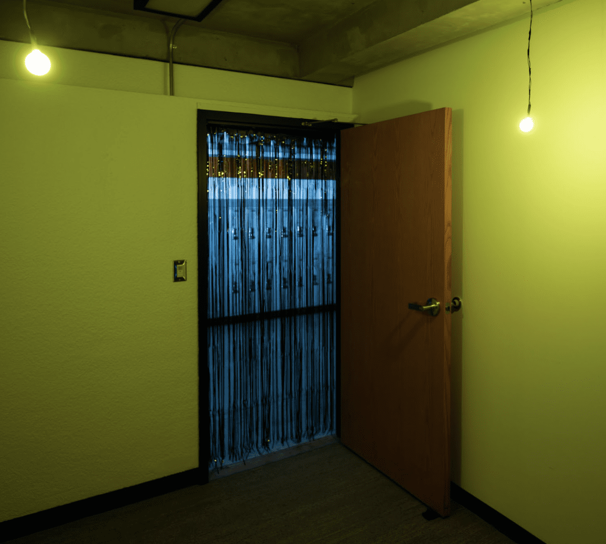 A small room bathed in yellow light. The doorway is covered in a curtain of tinsel strips. Two small lights hang low from the ceiling