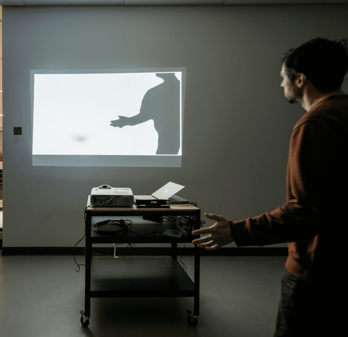 A small projection is displayed on a wall. The video shows a silhouette of a person, mirroring a person standing in the room.