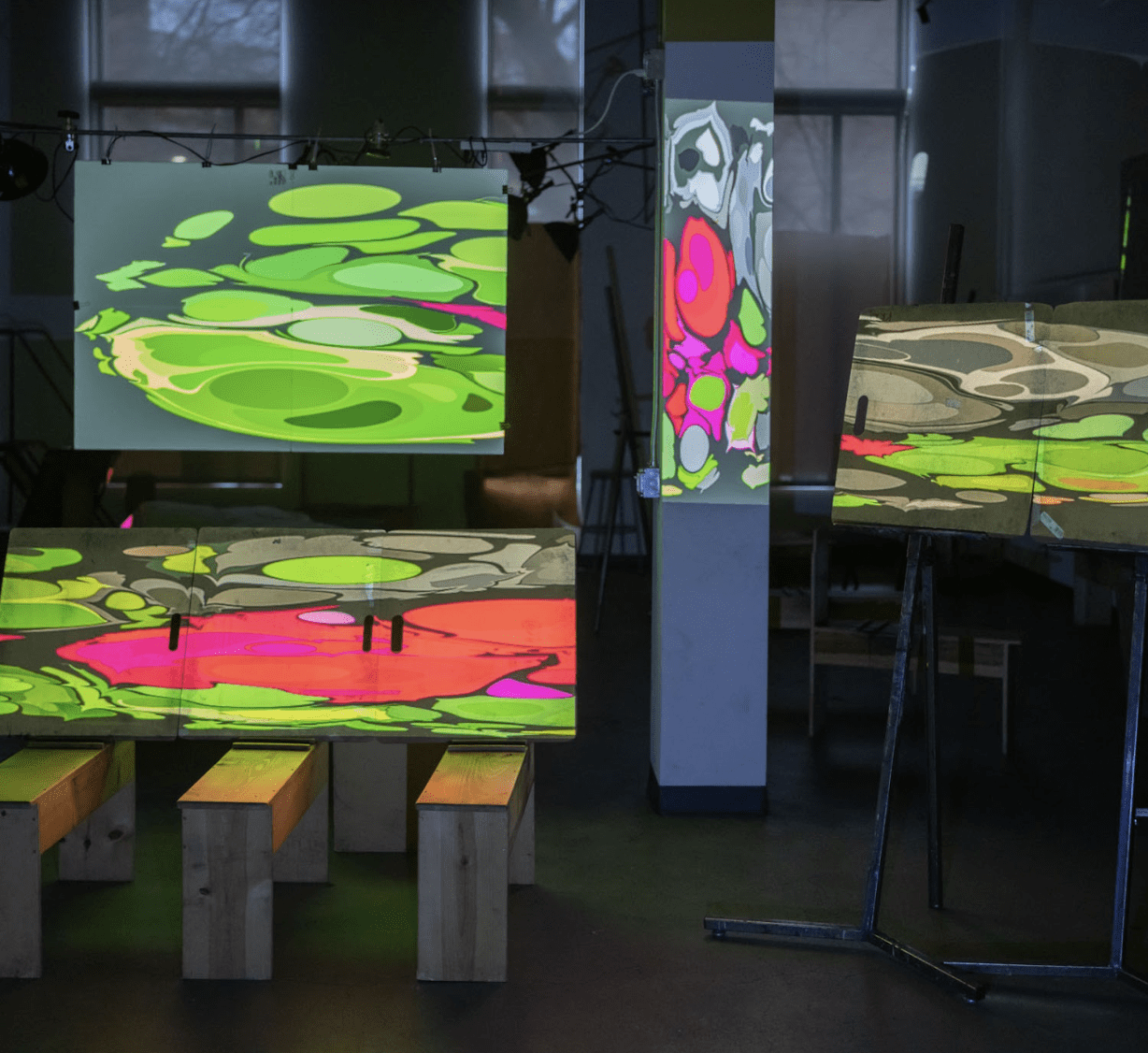 Several pedestals and benches are arranged in the room. Each stand holds a canvas. Videos of dripping color are projection mapped onto each of the canvases