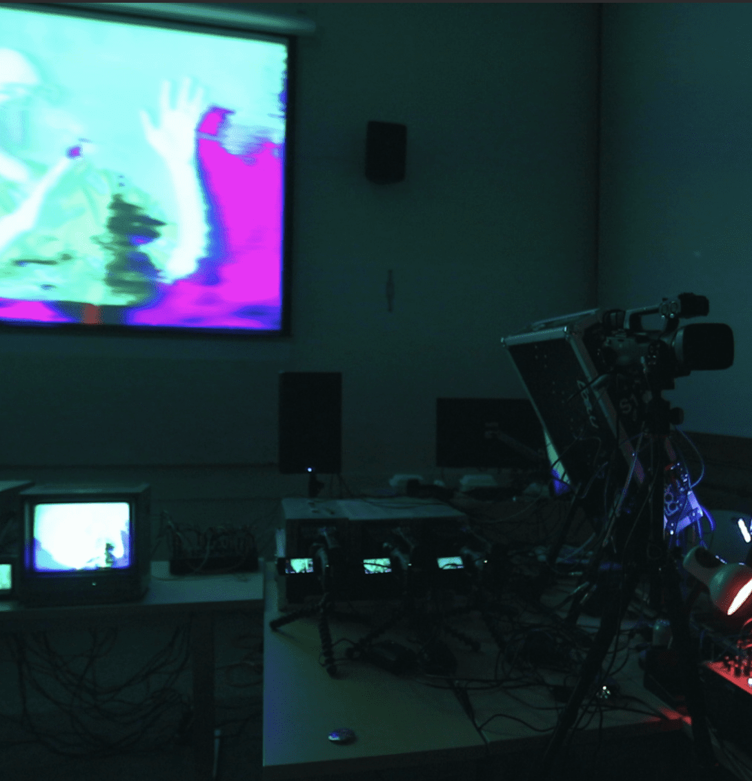 An abstracted video is displayed on a projector screen in the background. In the foreground is a camera and a tech setup filled with colorful wires.
