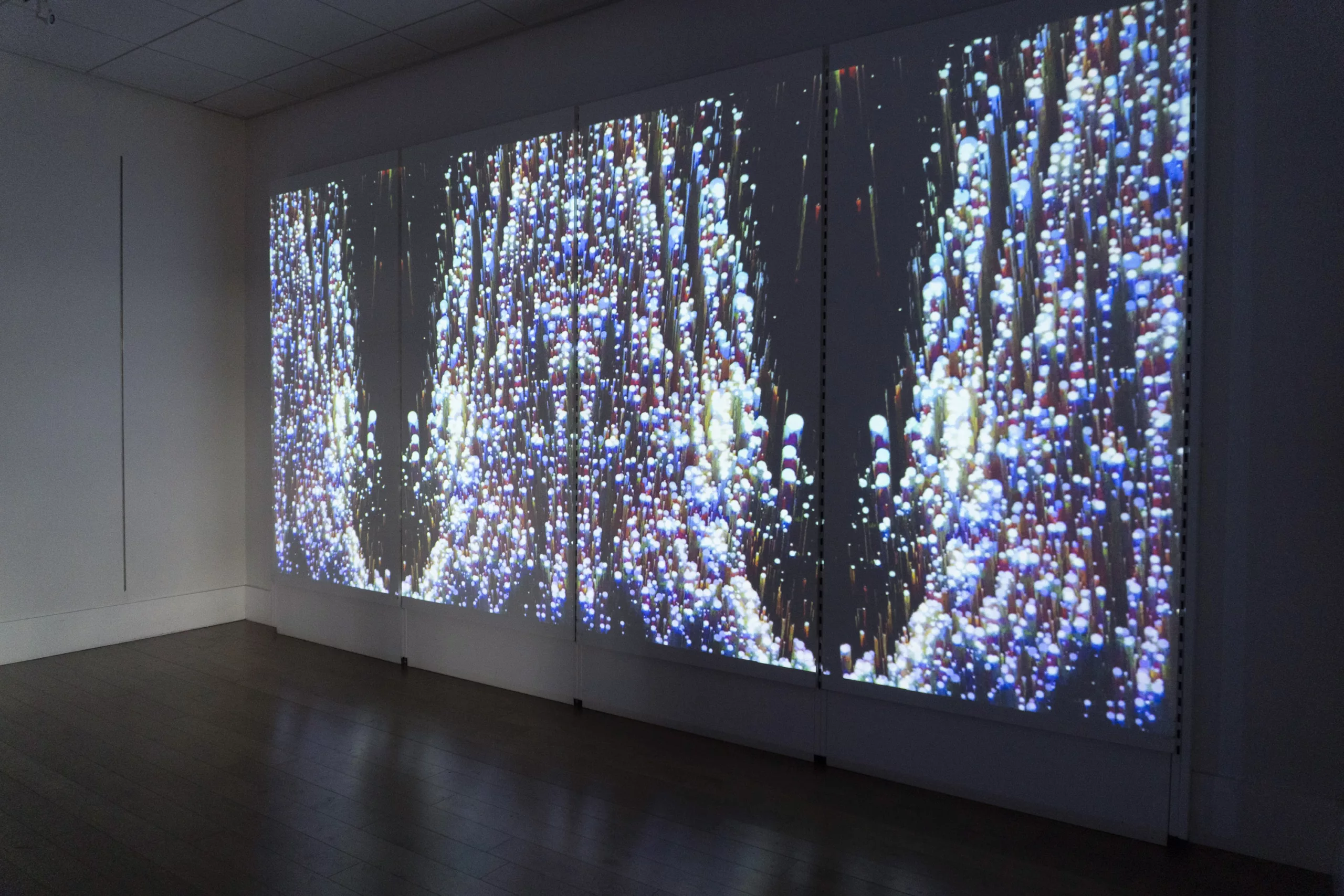 A video is projected onto a long wall. In the video, spherical balls of bright white, purple, and blue form patterns akin to waves of water.