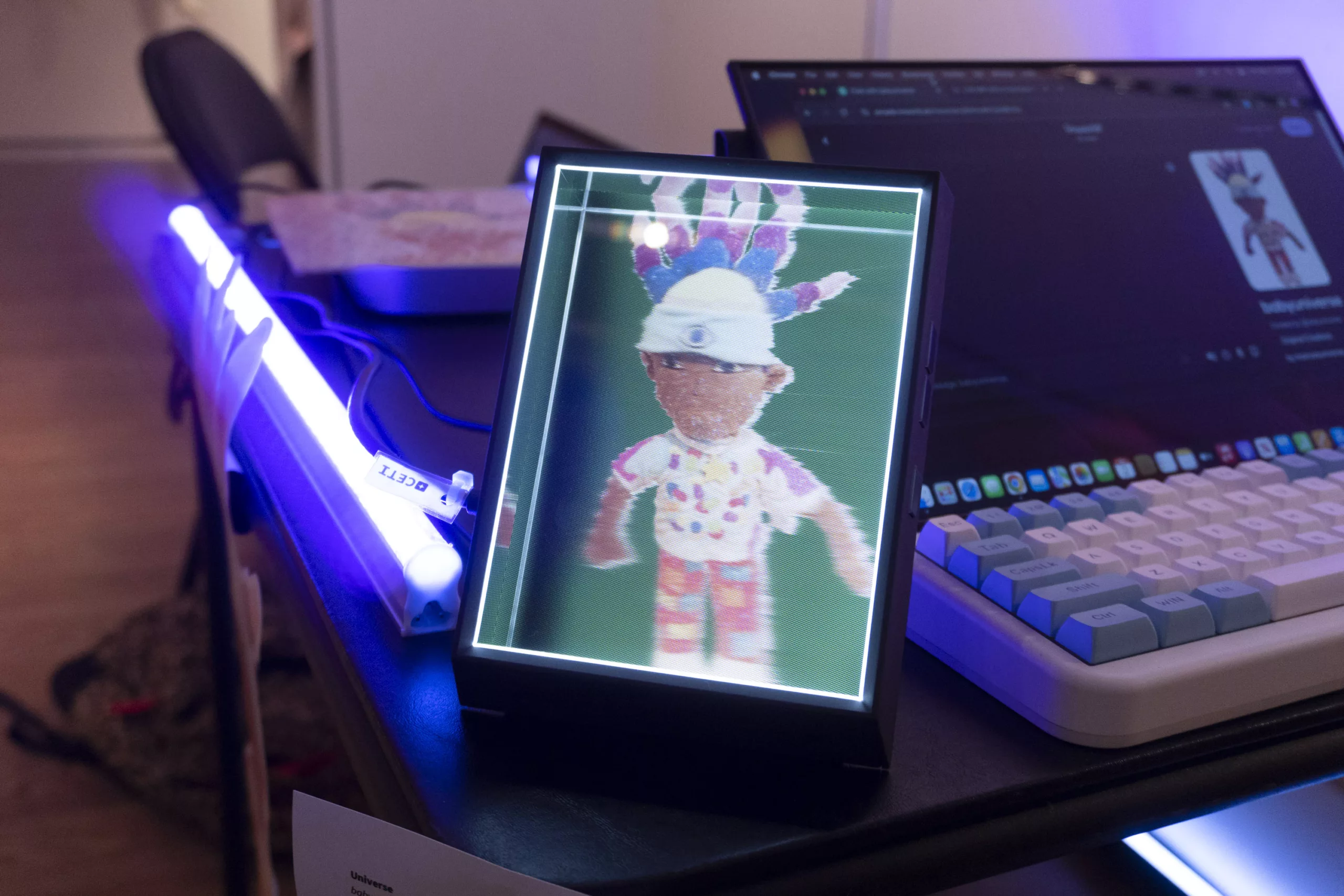 A hologram frame sits on a table next to a keyboard and computer. The image in the hologram frame is of a cloth doll with rainbow colored locs, a white beanie, and a brightly patterned outfit. On the computer screen is an ai chat program that allows visitors to speak to babyuniverse.
