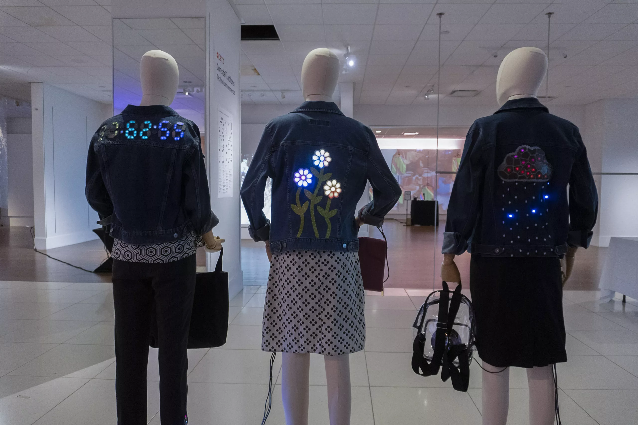 Three mannequins face away from the camera. The outfits are adorned with LED lights. The jacket on the mannequin to the left has numbers formatted like a clock timer, with colons between 3 sets of two digits. The mannequin in the middle has a jacket with glowing flowers. The mannequin on the right features a jacket with a rain cloud on the back. The cloud is embroidered and the rain drops are LED lights.