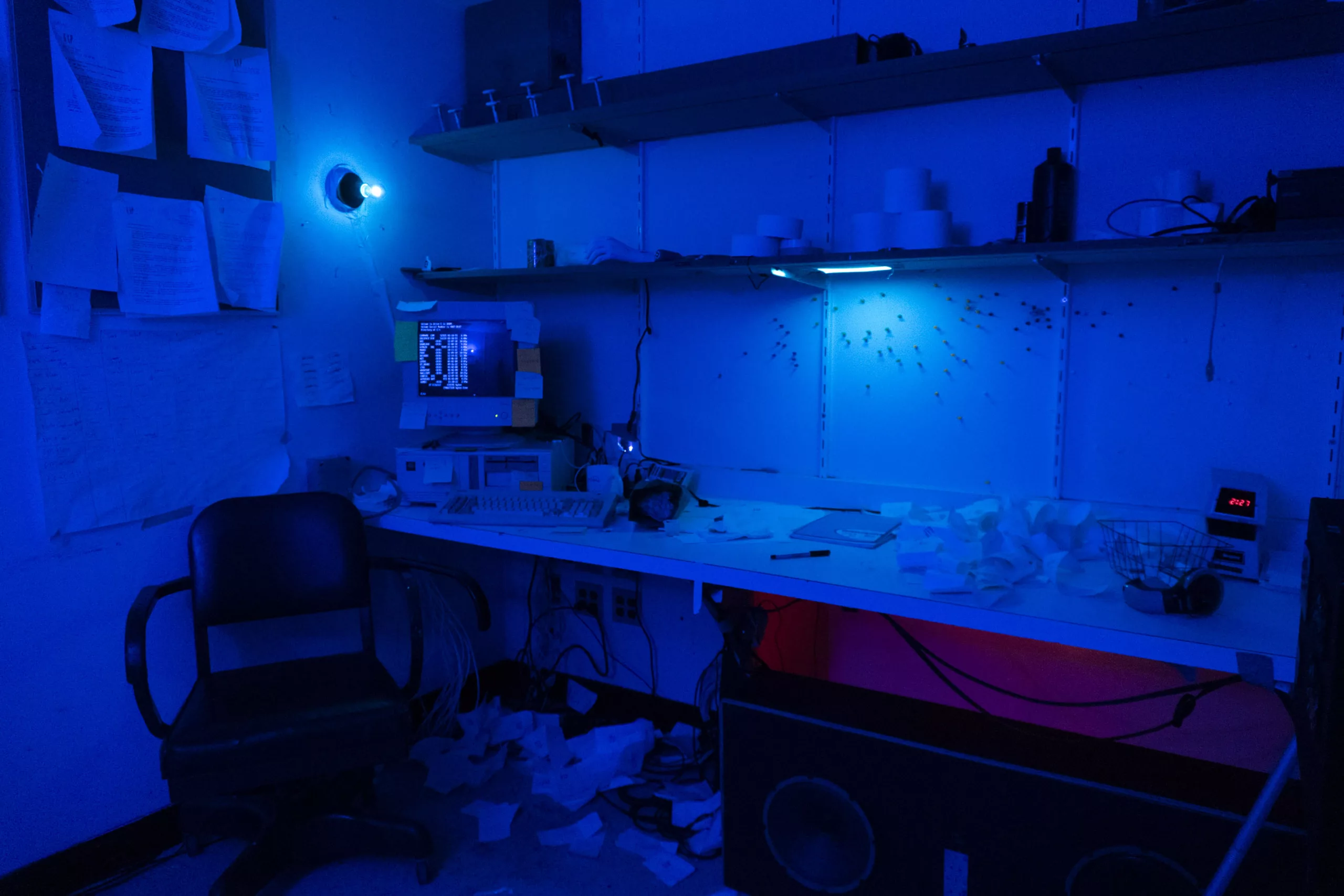 A messy managers office is bathed in supersaturated blue light. Papers are strewn everywhere - on the desk, on the floor, and on the walls. An old computer sits in the corner with the terminal open.