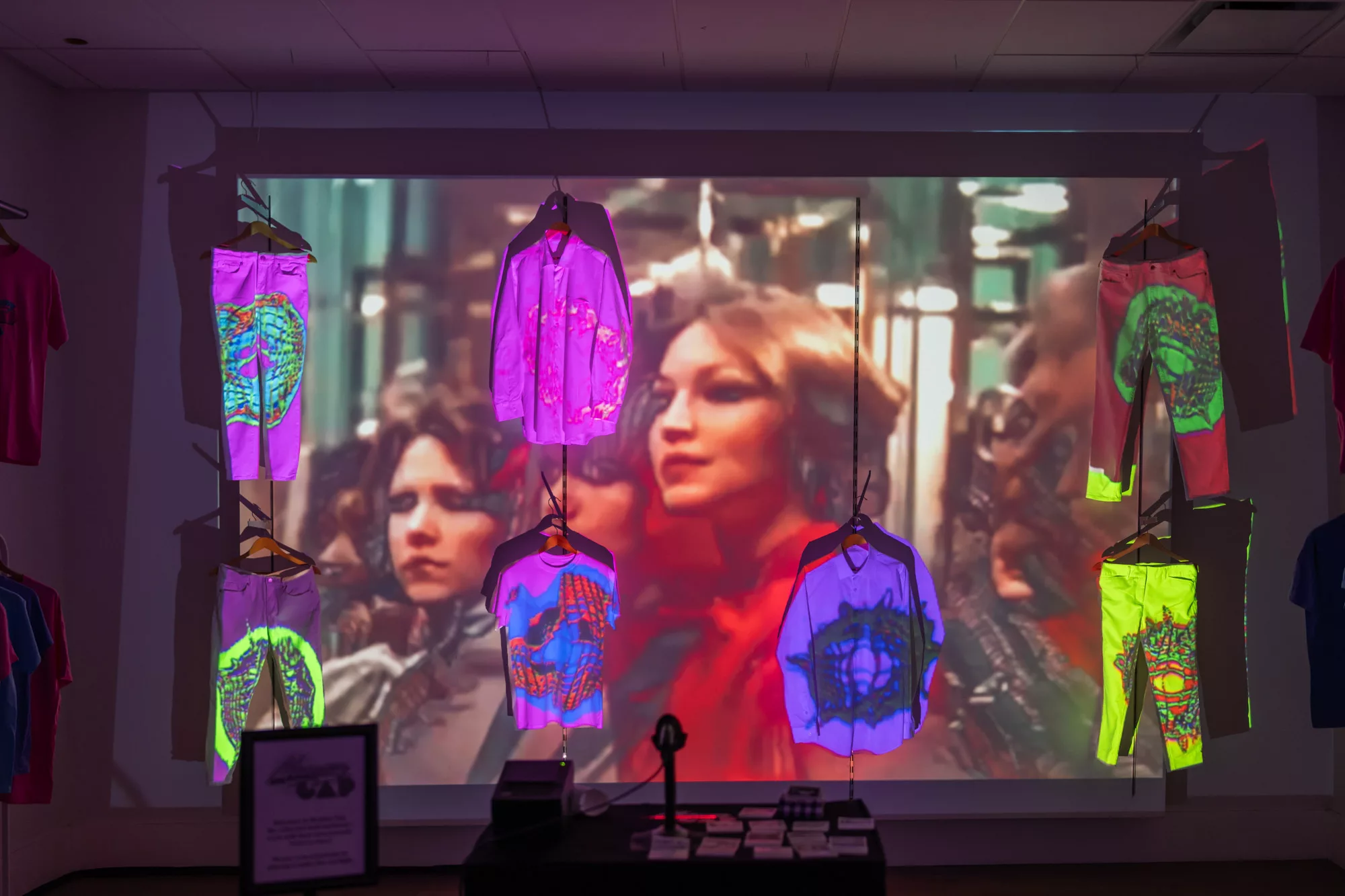A projected video shines on two rows of shirts and pants that are hung on a wall. Brightly colored, abstract shapes are projected onto the shirts and pants. In the negative space between the clothing, a second video is projected. The second video features two people in outfits indicative of the 80s. A table sits in front of the projection. On the table is a barcode scanner and a receipt printer. To the right of these, also on the table, are a grid of short receipts.