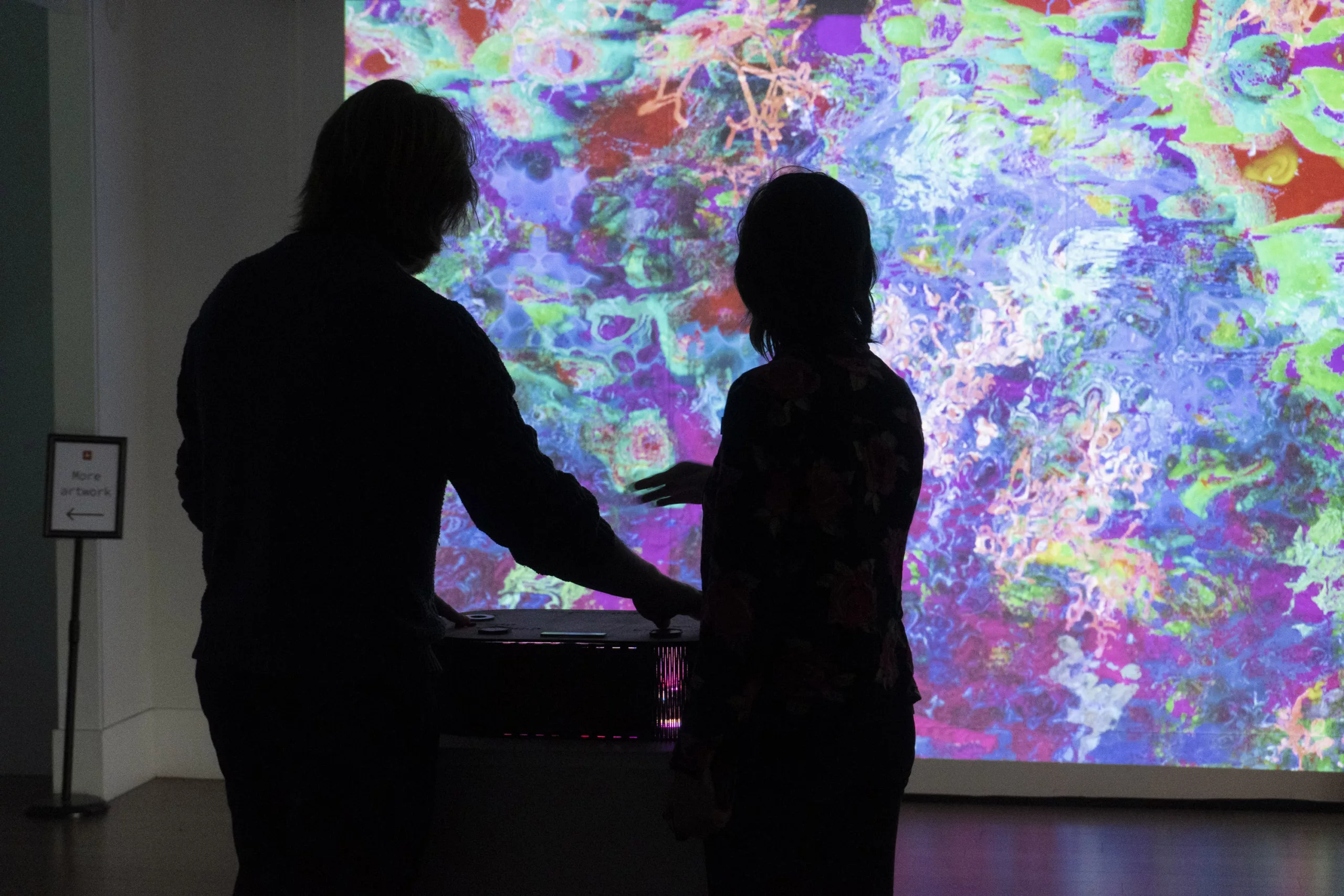 Abstract, swirling colors are projected onto a wall. The silhouettes of two people can be seen in front of the projection. They are standing by a rounded box with touch sensor controls.