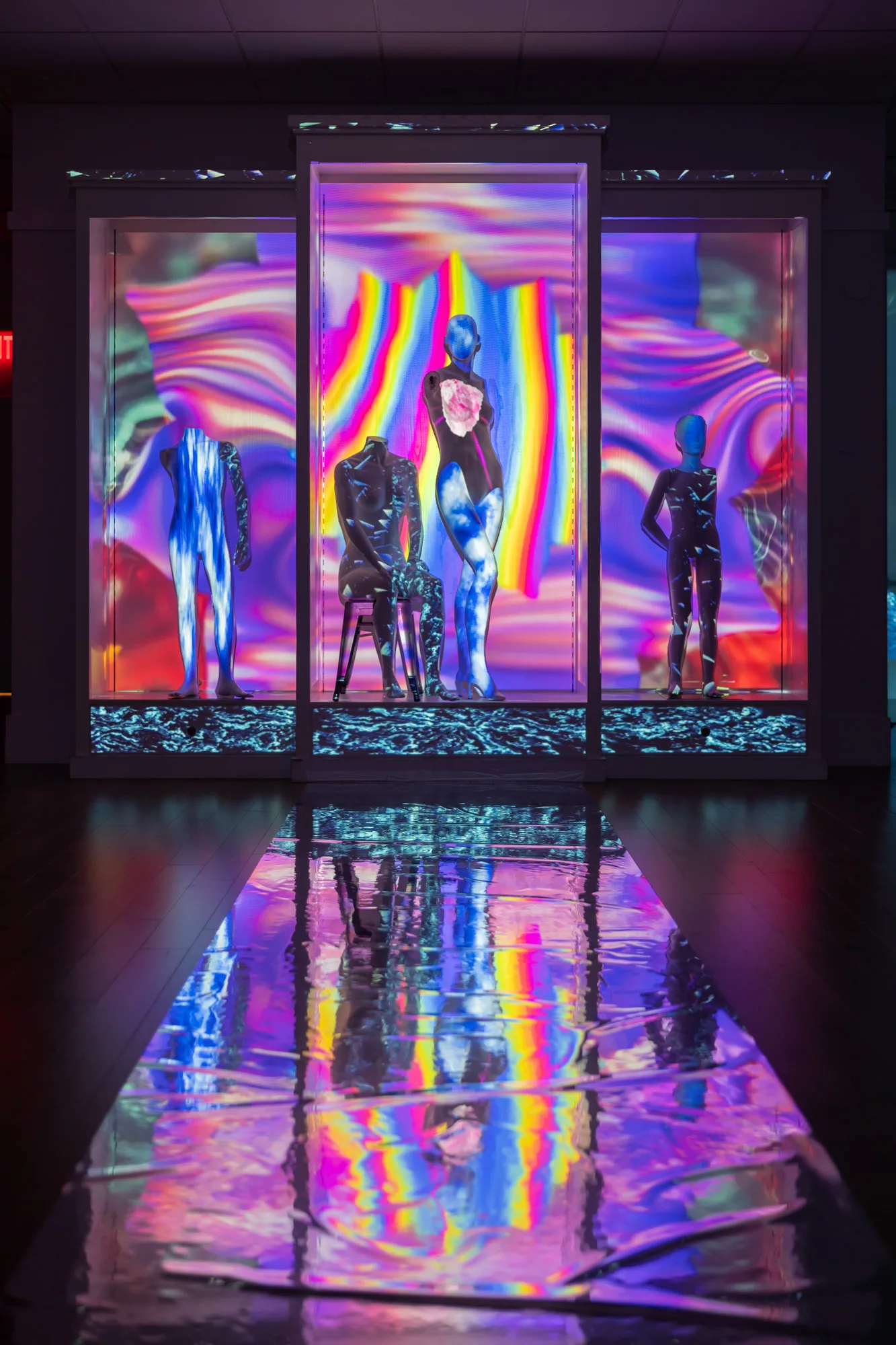 A display case, split into three sections. Four mannequins stand in the case - one in each of the side sections, and two (one standing and one sitting) are in the middle section. A shiny material is placed on the ground like a runway. A rainbow colored video is projection mapped onto the display case and mannequins.
