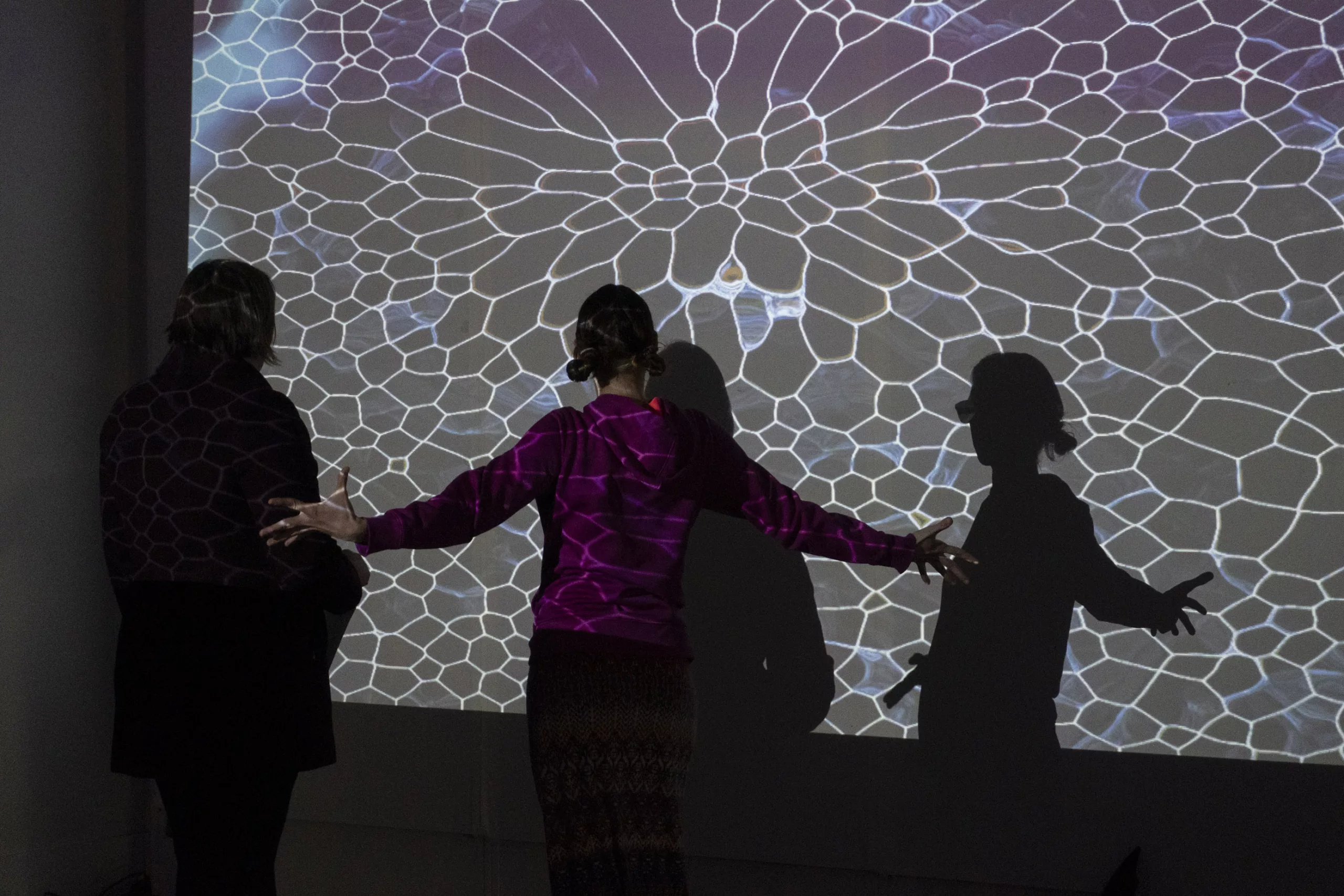 Two people stand with their backs to the camera, facing a projected video on a wall. The person near the center stretches their arms out wide. The video features an organic mesh web.