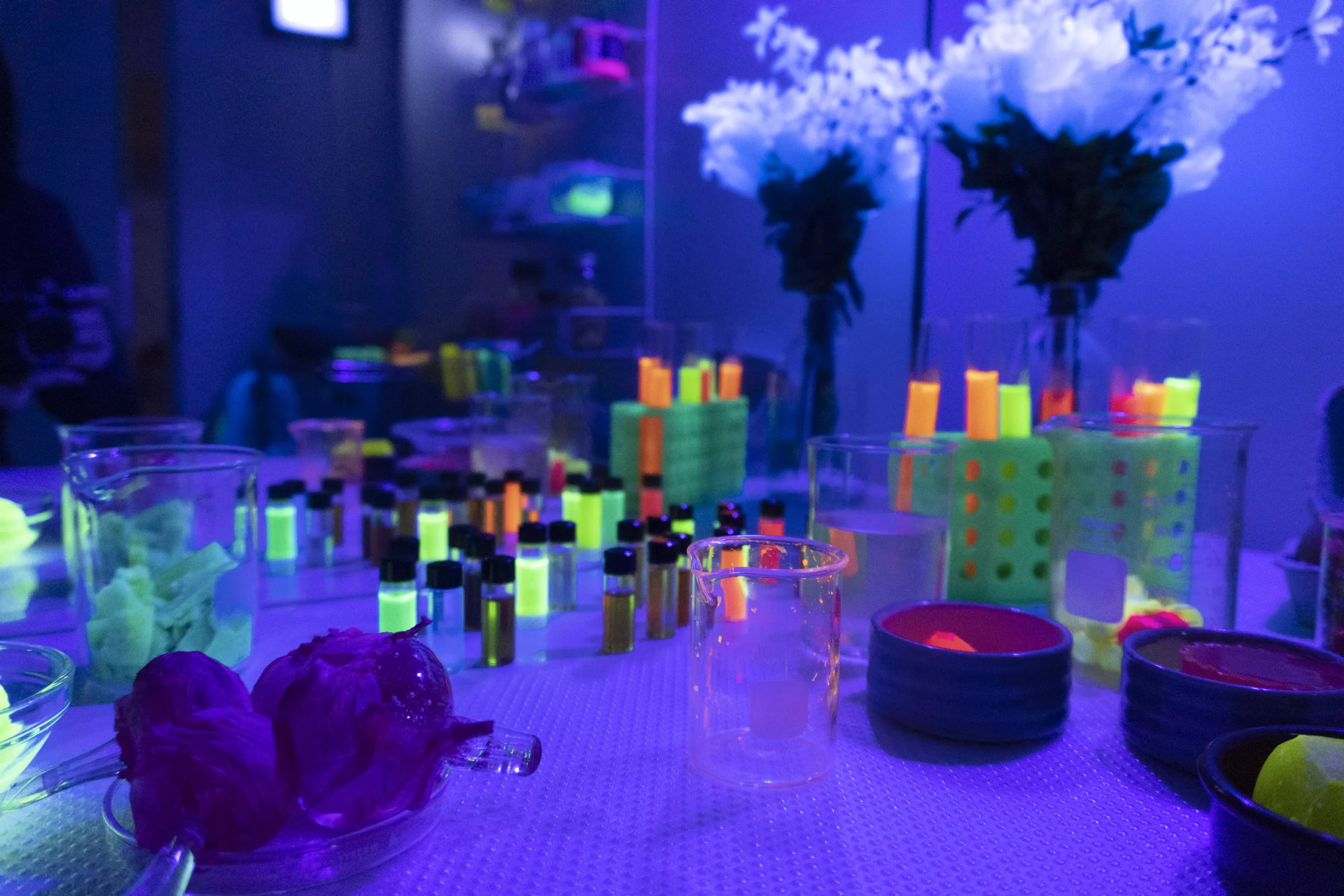 In a small room bathed in blue light, a table is set with a bouquet of glowing white flowers and a conglomeration of small glass cylinders. The glass containers are filled with glowing, UV reactive chemicals and dyes.