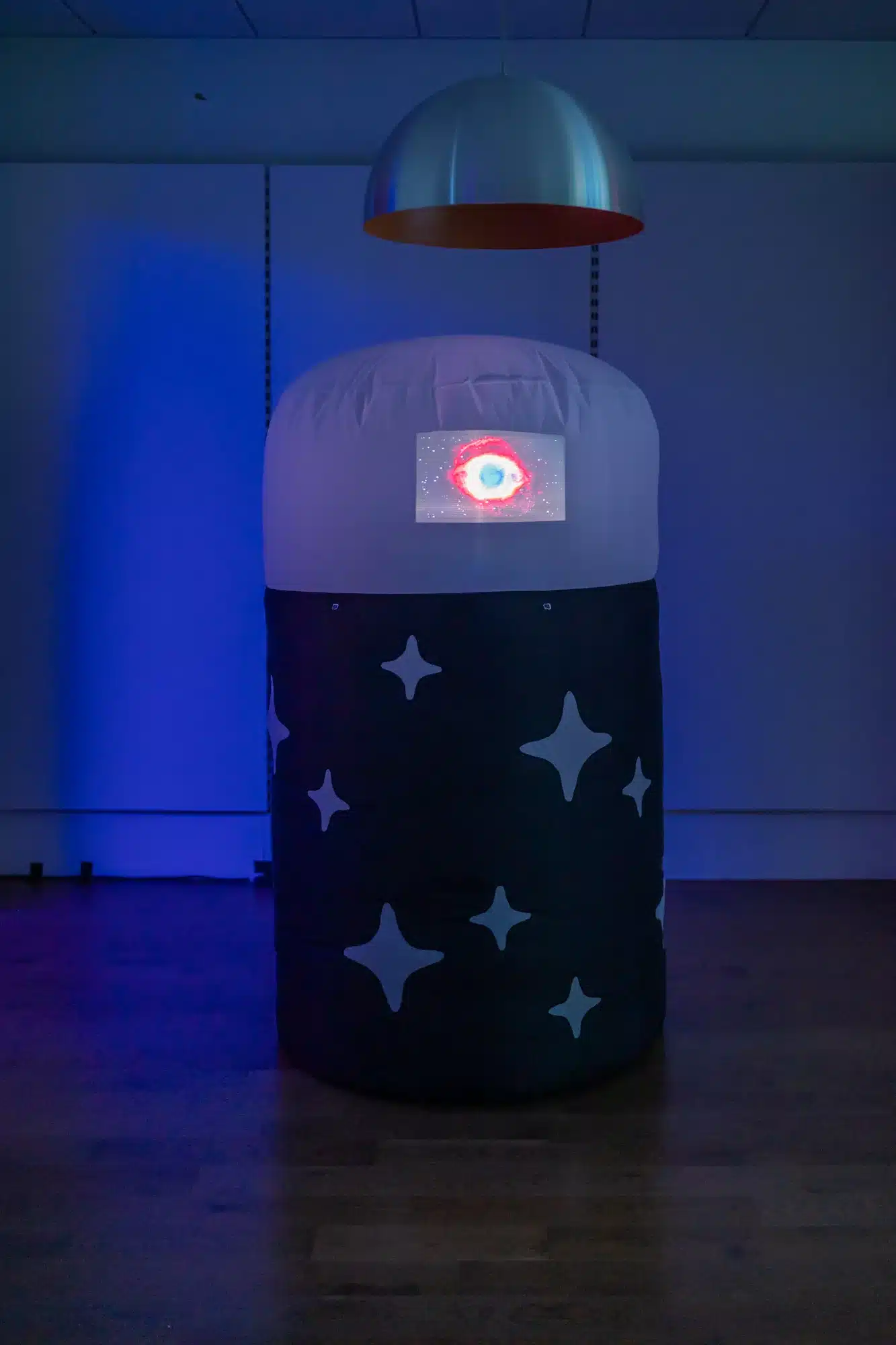 A large cylindrical structure stands in a room filled with blue light. The sculpture is formed using fabric that is inflated from the inside. The lower two thirds of the cylinder is black, scattered with stars of varying sizes. The top third is white. In the center of the top third, a video projected of a large, glowing eye.