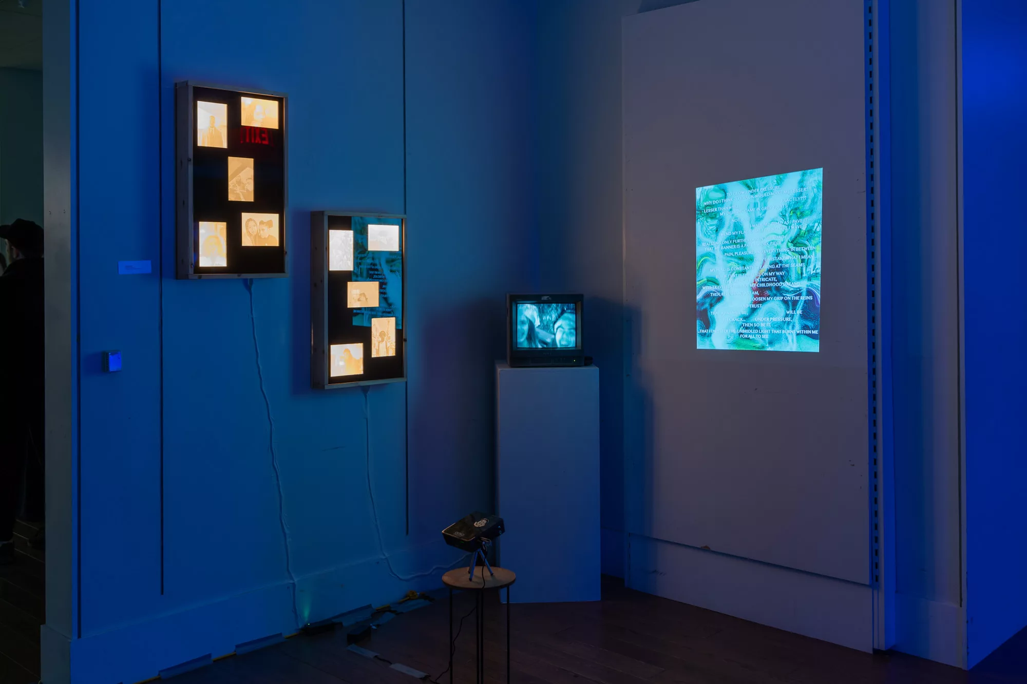 A video plays on a TV on a pedestal in the corner of a room. Two light boxes, each with 5 glowing, 3d printed photographs, hang on the wall to the left. A second video is projected on the the wall to the right of the pedestal.