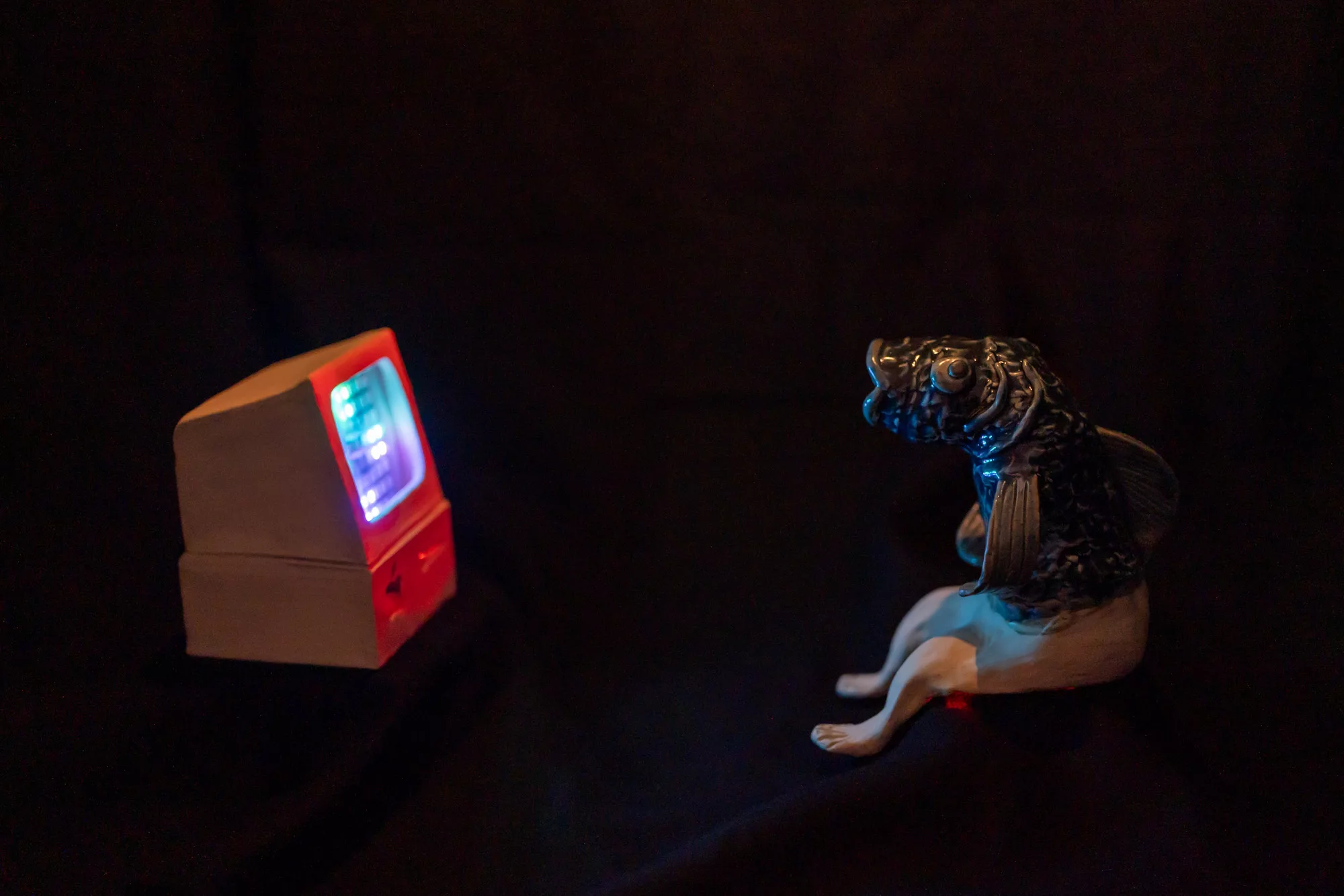 In a dark space, two ceramic sculptures face each other: a TV and an anthropomorphic figure with a fish head and human legs. LED lights shine from the TV screen.