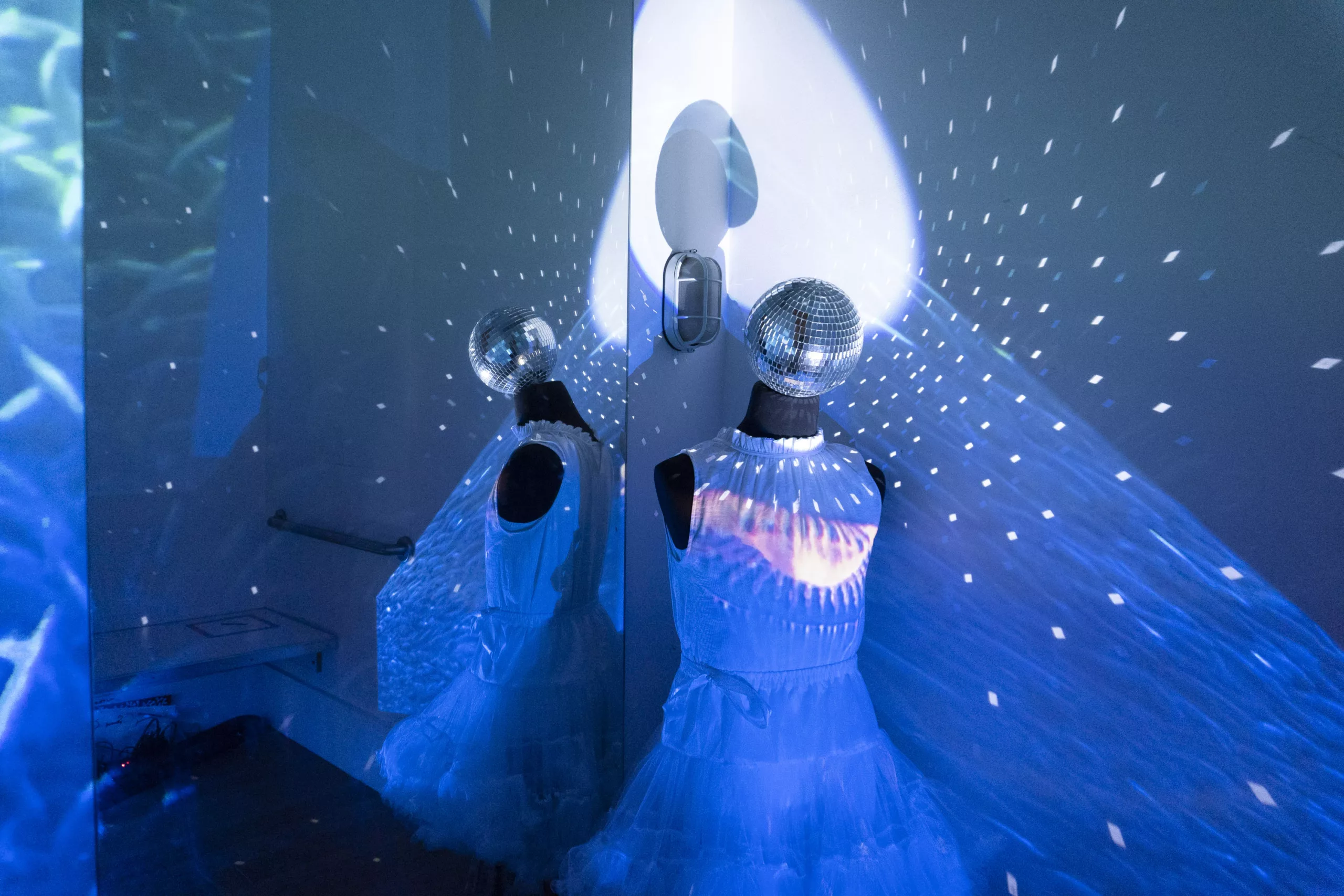 A mannequin in a flowy white dress faces away from the mirror of a fitting room. A sphere of mirrors acts as the mannequins face, reflecting light in all directions. The walls of the room are filled with projections of waves and sea life.